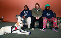 Article photo: four people sitting on a couch with a yellow Labrador in a red service vest lying on the floor in front of them