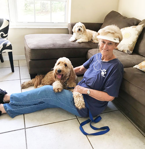 A woman in a white visor sitting on a tile floor beside a couch, with a cream colored curly coated dog on a blue leash resting across her legs and a small white dog lying on the couch behind.