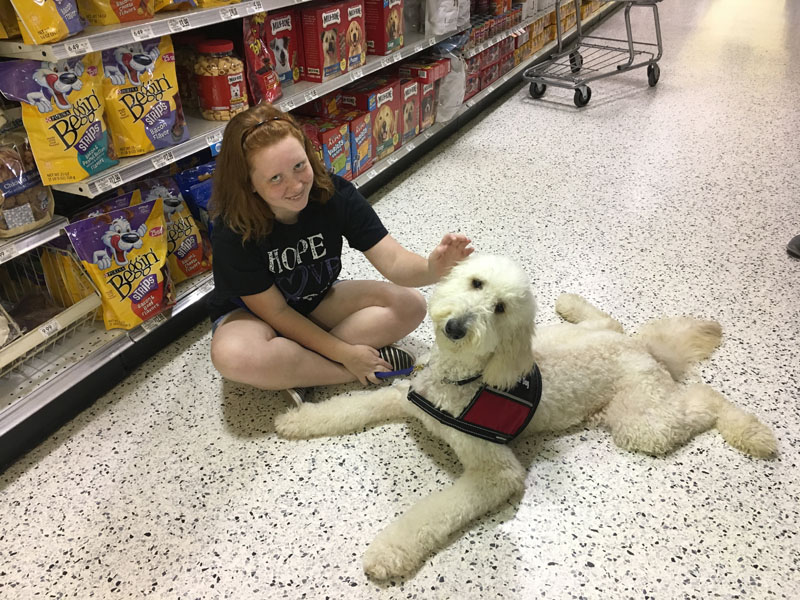 A girl sits cross legged on a supermarket pet food aisle floor with her hand on a large white curly coated dog lying beside her in a service dog vest with a red panel.