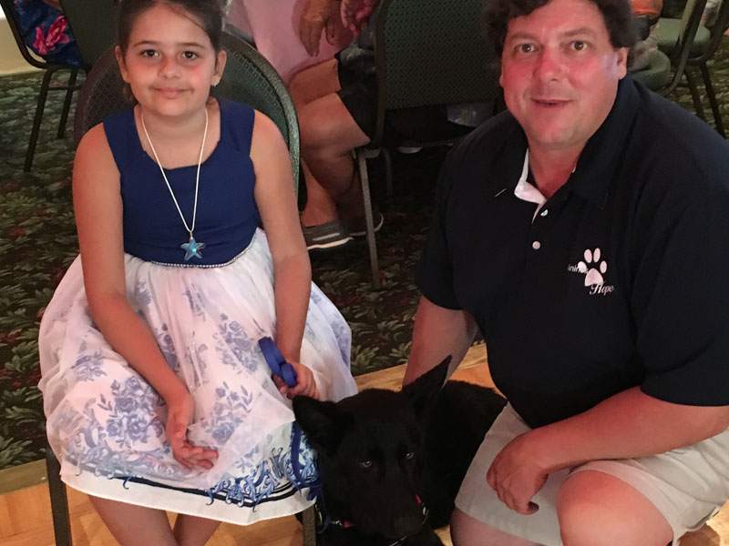 A girl in a blue and white dress seated on a chair at an indoor event, with a man in a Canines 4 Hope polo shirt kneeling beside a black German Shepherd in front of her.