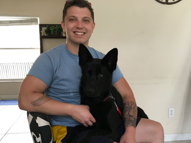 A young man in a light blue T-shirt sits indoors with a black German Shepherd puppy resting on his lap.