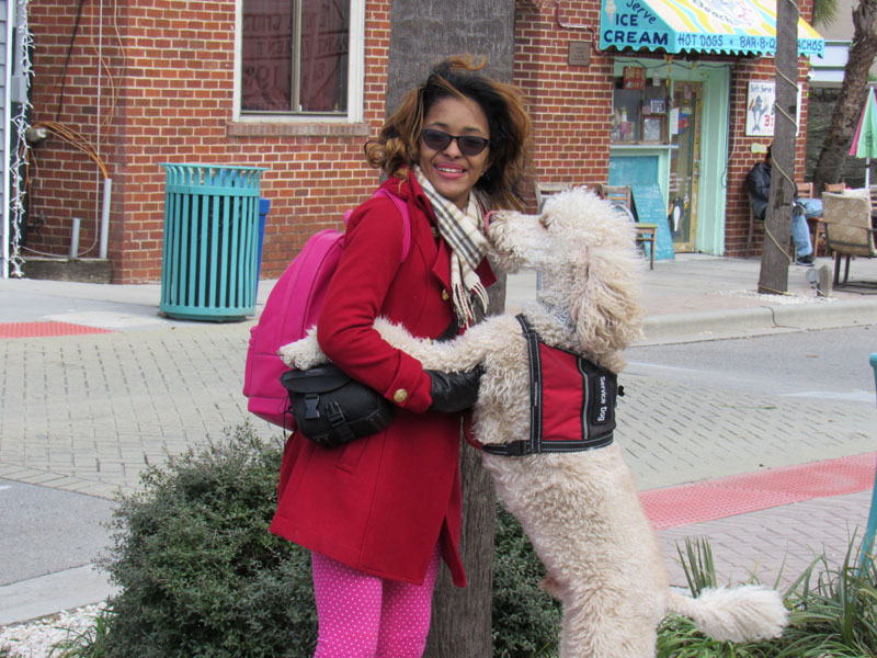 A woman in a red coat with a pink backpack stands on a sidewalk outside an ice cream shop while a large cream coloured curly coated dog in a red Service Dog vest rests its front paws in her hands.