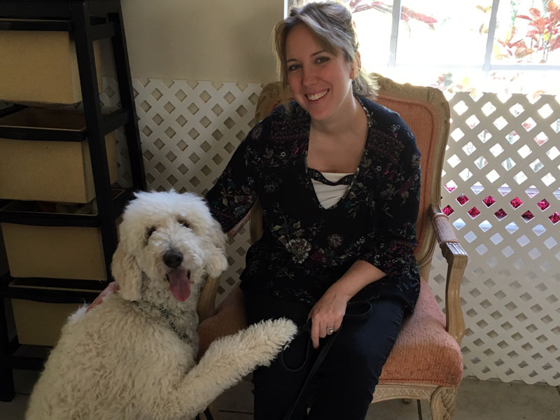 A woman sits in an armchair beside a large white curly coated dog that has rested one paw on her lap.