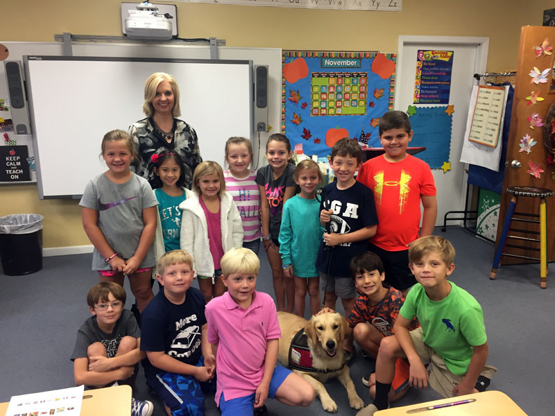 A woman and about a dozen young children gathered in a classroom around a yellow Labrador in a working vest, in front of a whiteboard and a November calendar board.