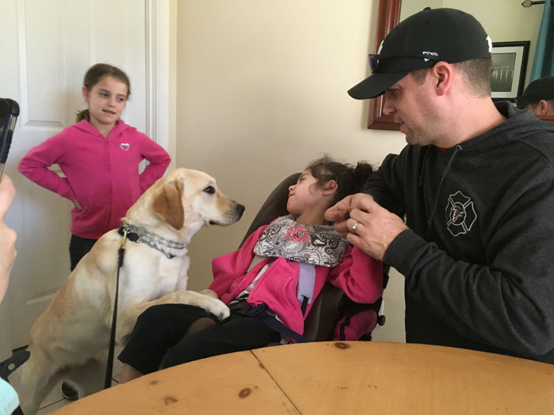A yellow Labrador sits beside a young girl in a padded high backed support chair at a wooden table, while a man in a black baseball cap seated at the table holds her hand and a girl in a pink hoodie stands in the doorway behind them.