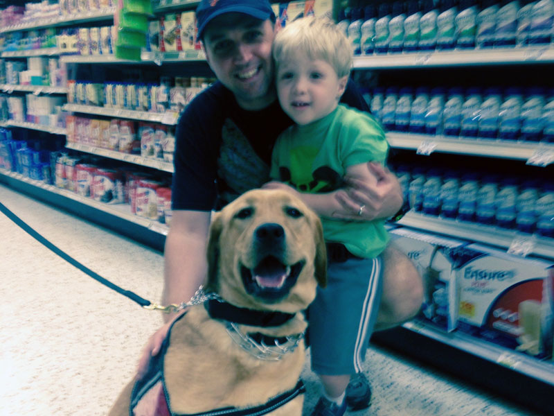 A man in a baseball cap crouching in a supermarket aisle with a small child on his knee and a yellow Labrador in a harness sitting in front of them.