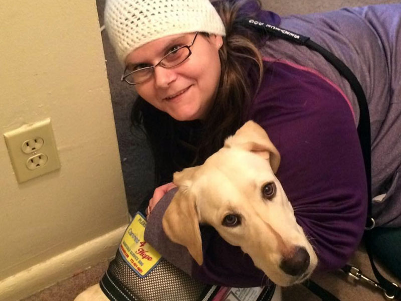 A woman in a white knitted hat kneeling in a hallway with her arms around a yellow Labrador wearing a Canines 4 Hope service dog vest.
