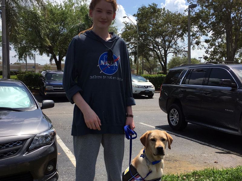 A teenager in a NASA sweatshirt stands in a parking lot holding a blue leash, with a yellow Labrador in a dark service vest sitting at their side.