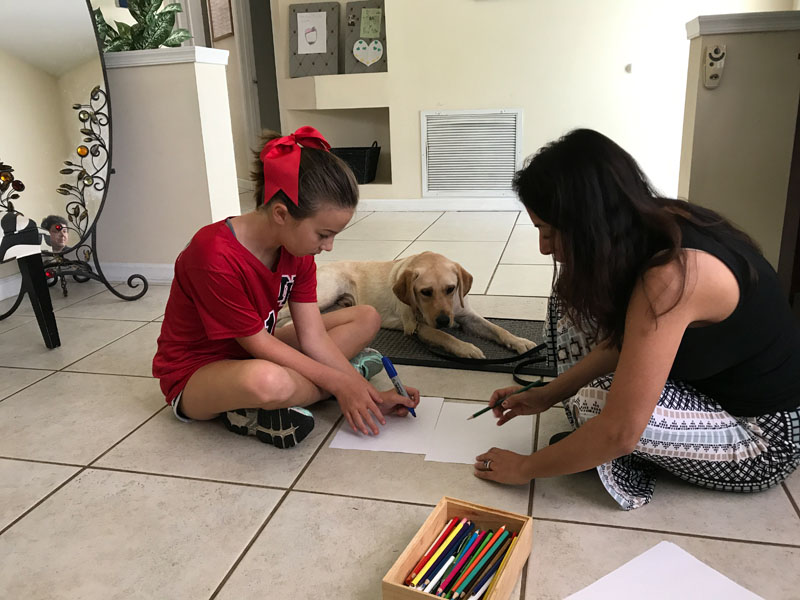 A girl in a red shirt and a woman kneel on a tile floor writing on sheets of paper, a box of coloured pencils beside them, while a yellow Labrador lies on a raised mat behind them.