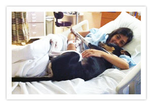 A person lying in a raised hospital bed rests a hand on a large black dog stretched out beside them on the blanket.